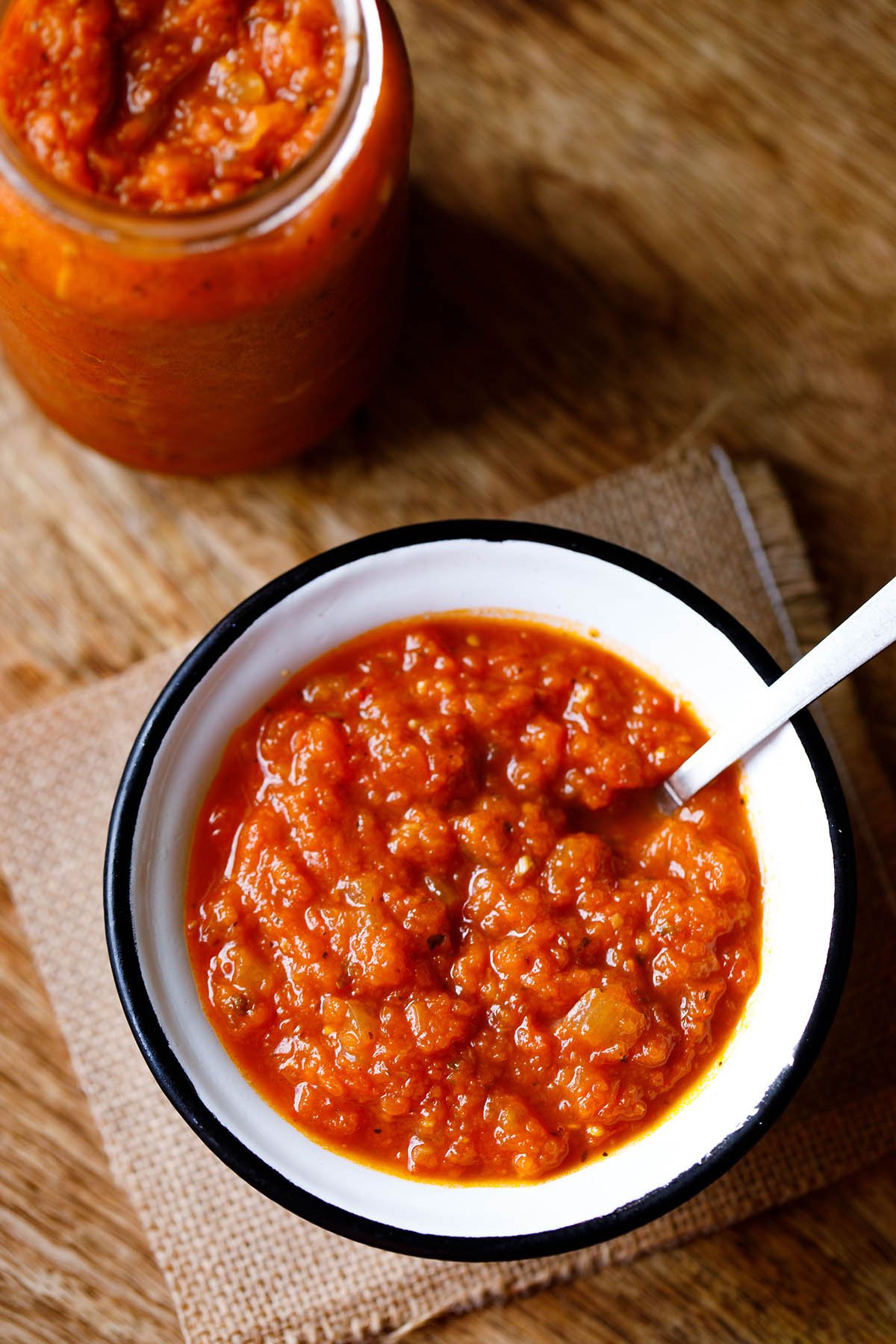 Glass jar filled with homemade marinara next to a bowl with a silver spoonful of marinara.