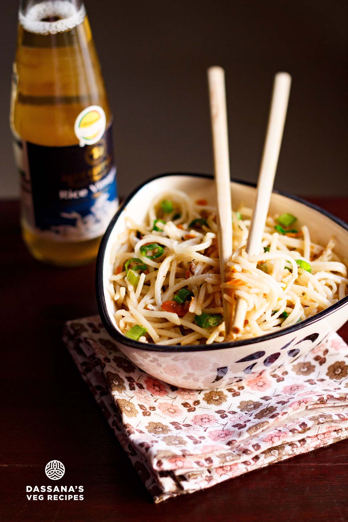 Hakka noodles served in a triangular bowl on a printed napkin with cream-colored bamboo chopsticks placed on top.