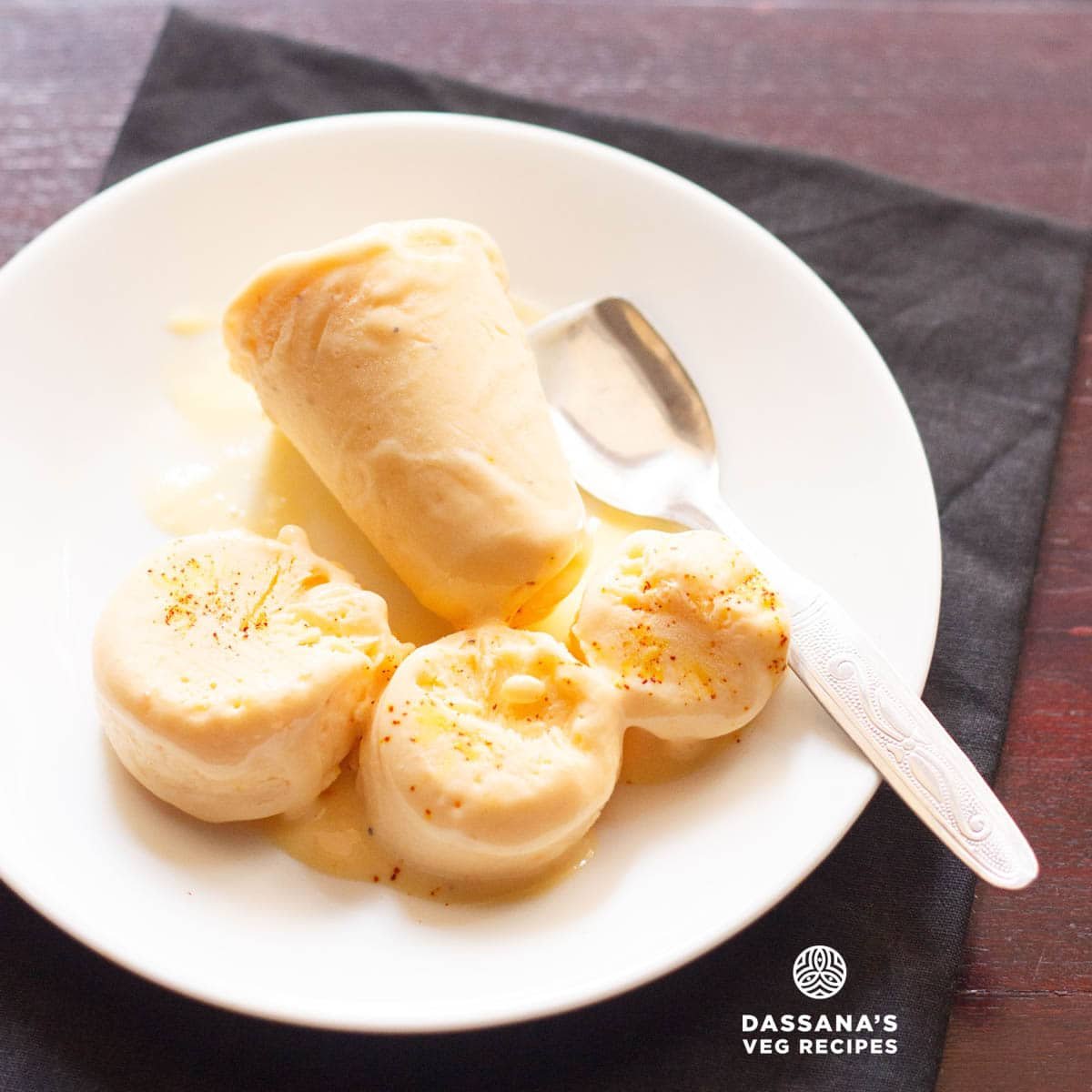 Mango kulfi sliced and whole served on a white plate with a steel spoon.