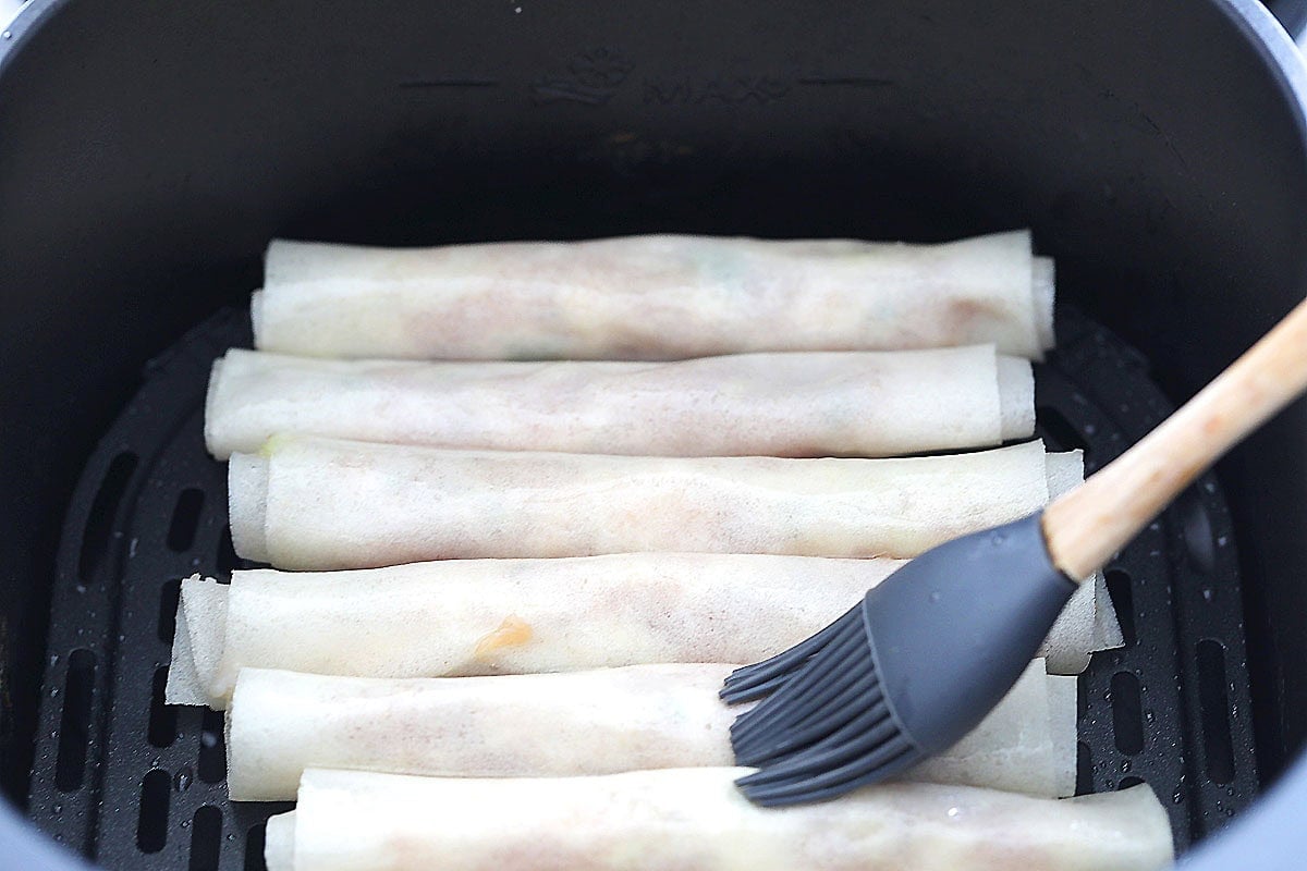 Uncooked lumpia in an air fryer basket, each roll lightly coated with oil.