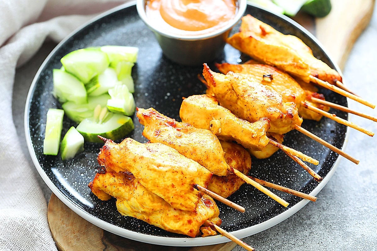 Golden-brown air fryer Thai chicken satay skewers with cucumber slices and Thai peanut satay dipping sauce on the side.